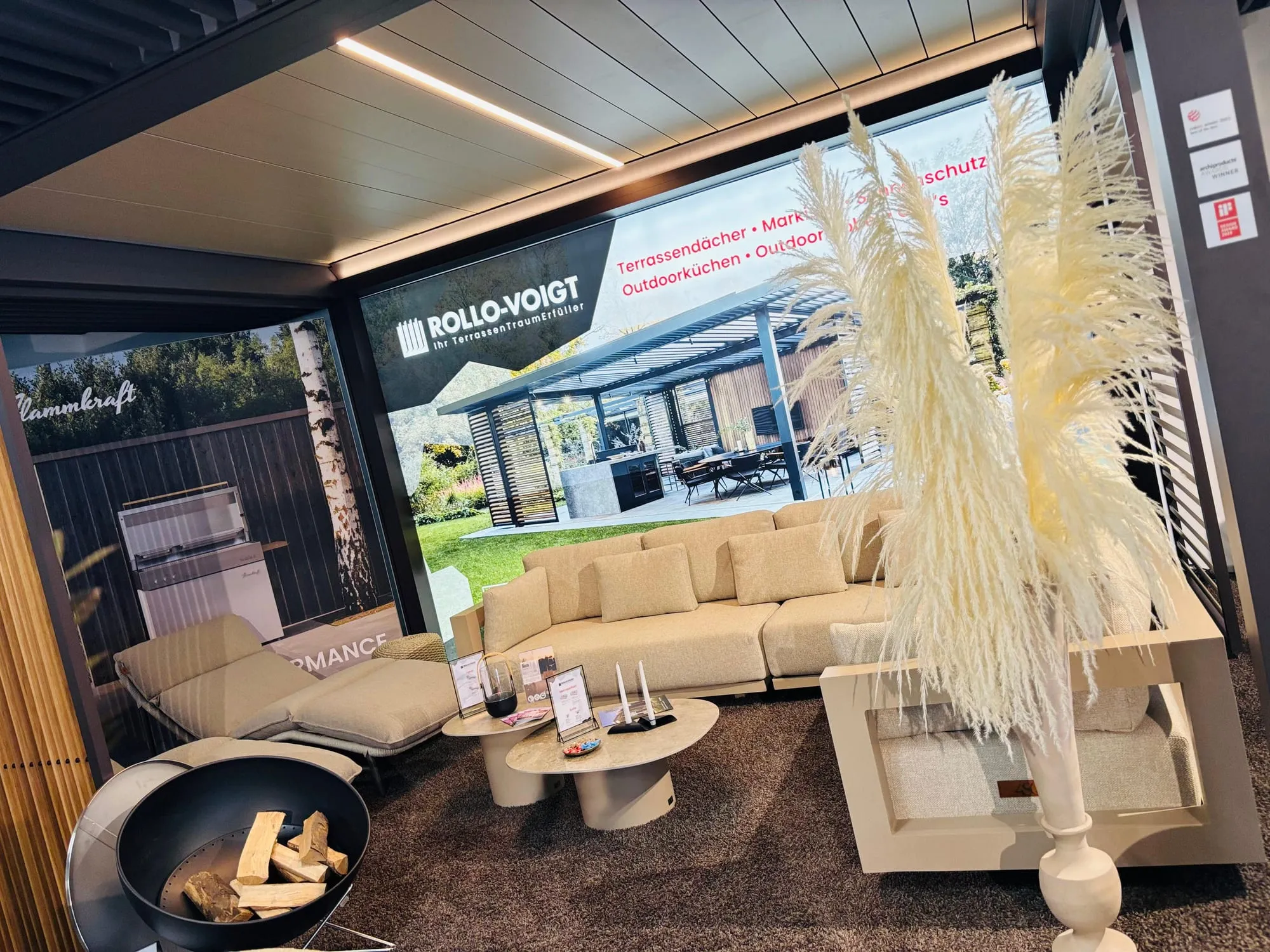 Lounge-Ecke im Pop-up-Store mit Sofa, Feuerschale und Pampasgras vor großformatiger Rollo-Voigt-Wand
